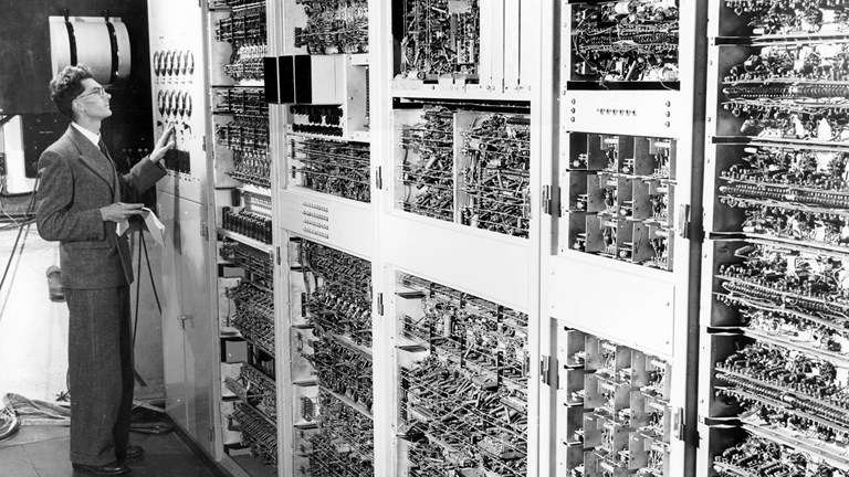 Black and white photo of a man in a suit operating a very old computer which is several cabinets in a row with many wires, switches and dials. 