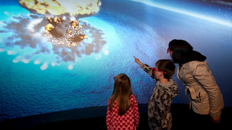 A woman and 2 children pointing at a large screen