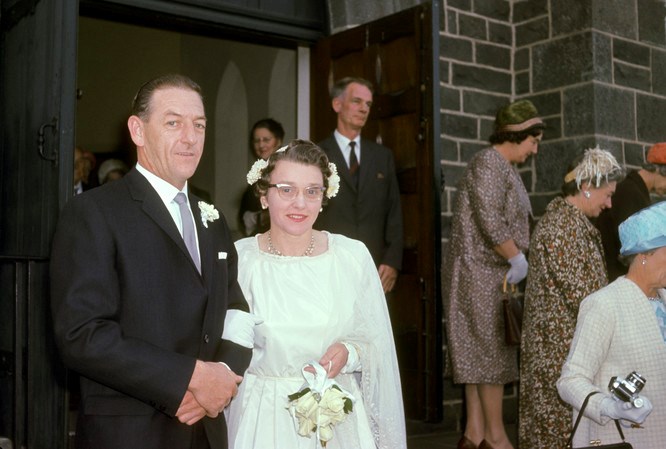 Bride and groom outside church to the left of image and facing camera. Some guests in background outside and inside the church.