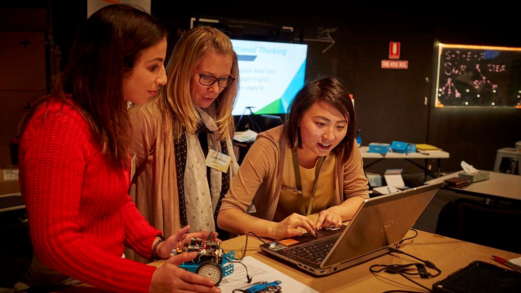 Two female teachers look on as a female museum staff member uses the mouse pad on a laptop. One teacher looks down at a blue car shaped robot with electronics on top.