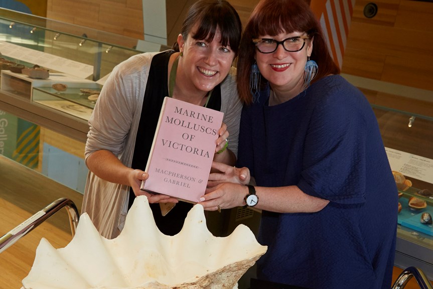 Two women hold a pink book in front of a large shell. 