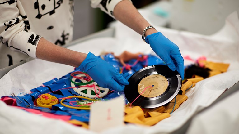 Conservator, wearing blue gloves, handling Jenny Bannister's blue plastic 'Je Suis Mod' dress, holding a vinyl record decoration.