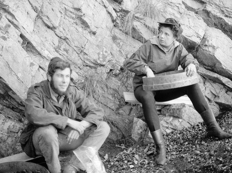 a black and white photo of a woman and a man sitting down and sifting soil