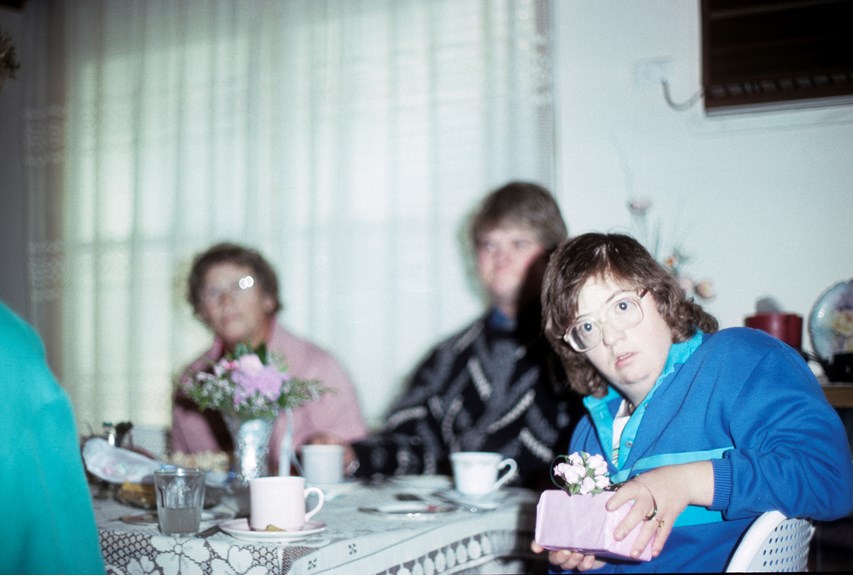 In foreground female wearing blue striped top holding a gift sitting at table set for a meal including a vase of pink flowers, with a male and female out of focus sitting in background at the table.