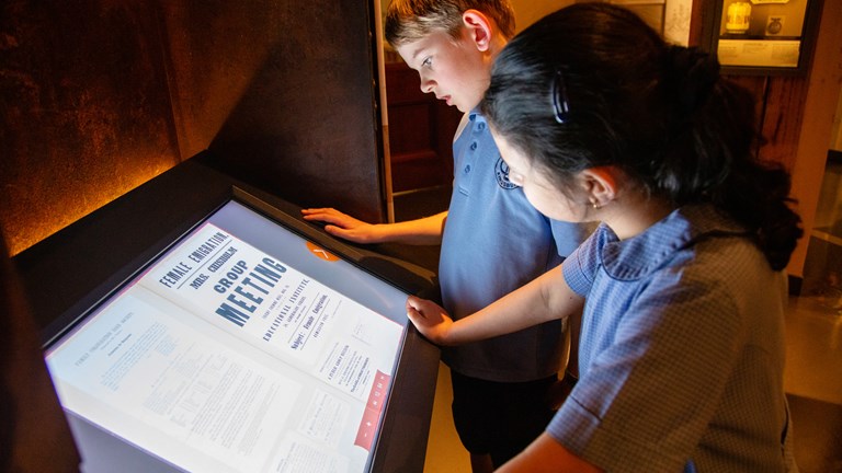 A boy with blonde hair and a girl with black hair looking at a digital screen in a museum gallery. 