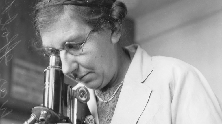 a black and white photo of a woman wearing glasses and a white coat, looking into a microscope