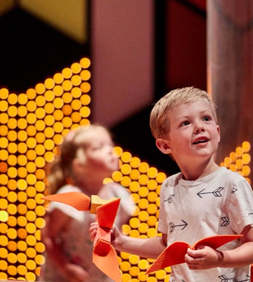 A boy holding a paper plane in a bright exhibition space. There is another child out of focus in the background.