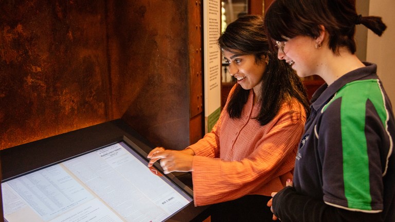 A woman with black hair and a young girl with brown hair and glasses look and smile at a digital screen. 