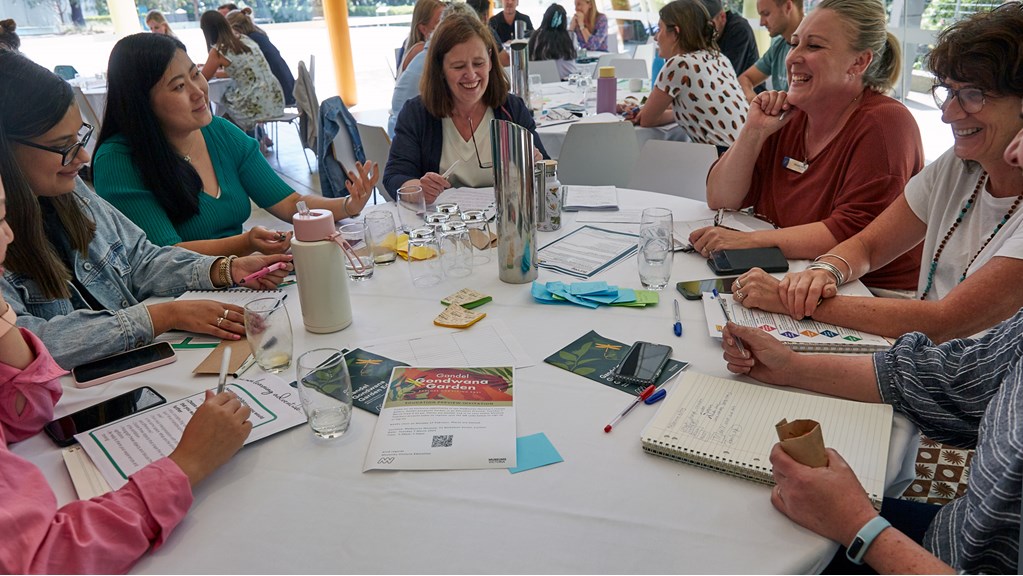 Group of women sitting at a round table with a white tablecloth. They have notepads and pens. There are museum leaflets on the table.