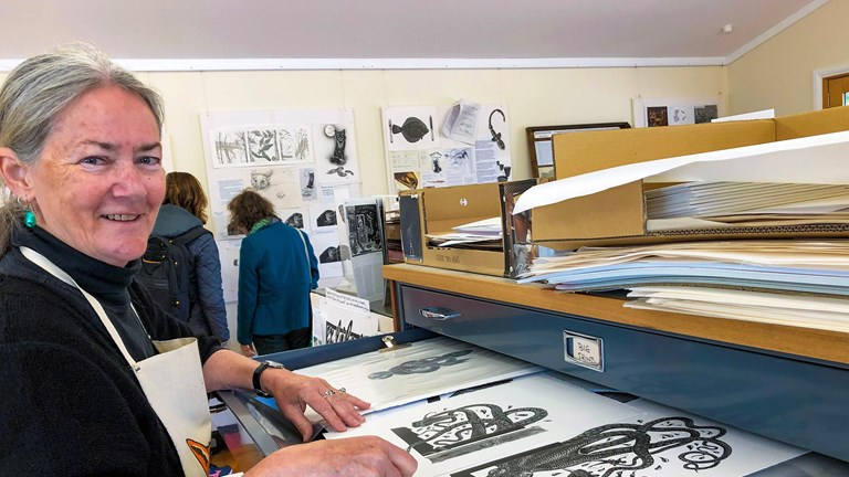 a photograph of a silver haired woman wearing an apron in front of a large drawer filled with black and white artistic prints