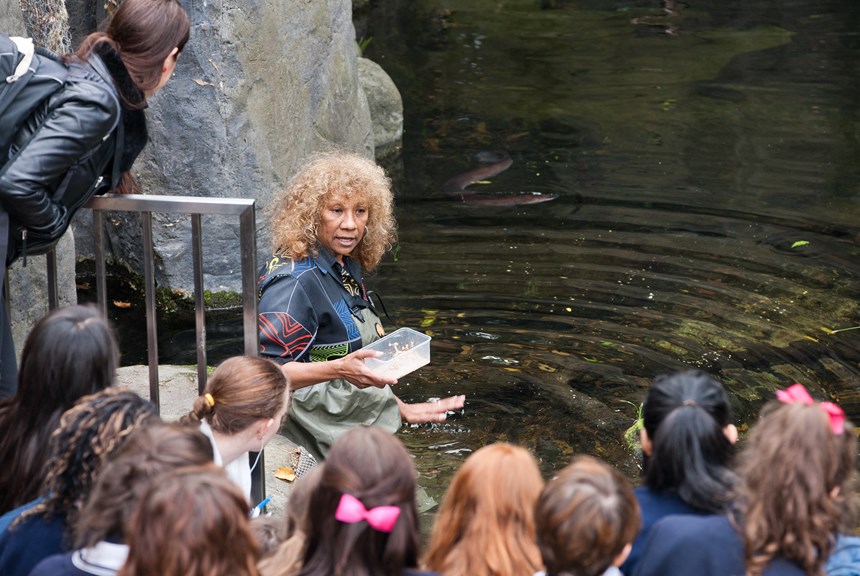 a woman gestures to a pool of water in front of schoolchildren