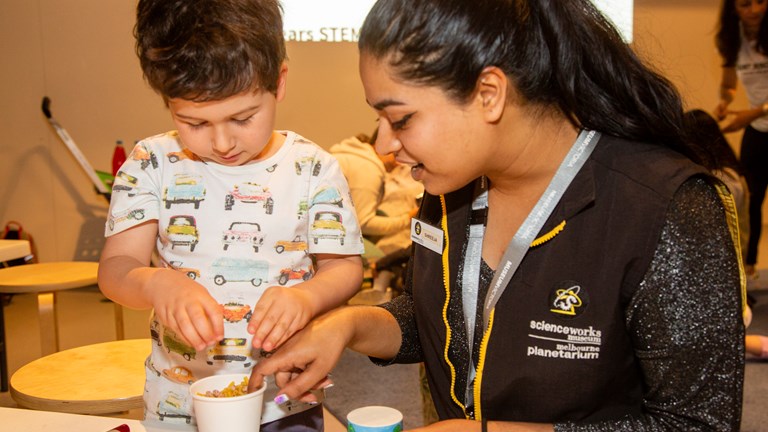 A woman with black hair in a ponytail sitting at a work table with a young boy with brown hair looking into a white cup full of things to create sound