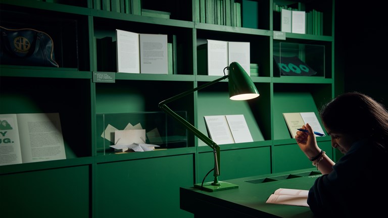 A girl sitting at a desk with pen in hand ready to write. The room is painted green and the walls are filled with objects and books. 