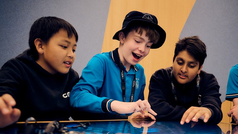 Three primary school students in school uniforms sit at a digital touch table. They excitedly touch the screen in front of them as they create a road safety ad in the Road to Zero Learning Studio at Melbourne Museum.