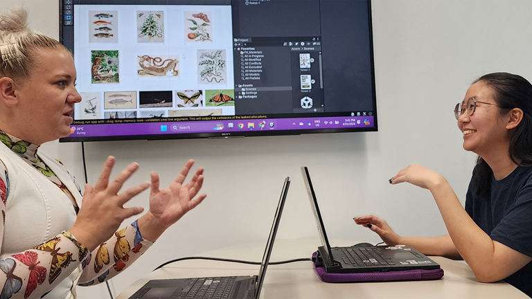 Michelle Chen and Caitlin Kozman chat about game design with a Unity scene builder screen mounted to the wall in the background.