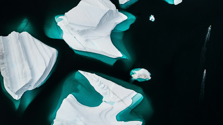 Aerial image of icebergs floating in a blue sea. Tiny boats are seen motoring past. 